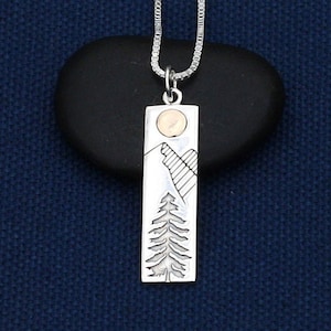 May include: A silver necklace with a rectangular pendant featuring a stylized mountain scene with a tree and a small gold circle.