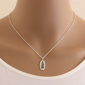 Op de afbeelding: Een zilveren ketting met een klein ovaal hanger met een eenvoudige silhouet van een dennenboom.