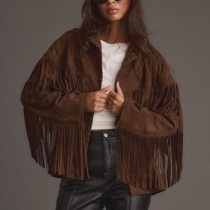 May include: A brown suede jacket with long fringe detailing on the sleeves and chest. The jacket is worn over a white top and paired with black leather trousers. The model is wearing black sunglasses.