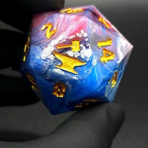 May include: A twenty-sided die featuring a swirling blend of blue, pink, and white resin with embedded silver leaf accents. The numbers are engraved in a bold font and filled with a metallic gold finish, including a hammer icon on the twenty face.
