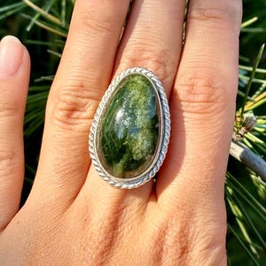 May include: A teardrop-shaped ring featuring a polished, mottled dark green stone set in a silver bezel. The stone displays natural variations in color, ranging from deep forest green to lighter mossy green tones. The setting is framed by a twisted rope-style silver border, creating a textured edge around the smooth, rounded surface of the gemstone.