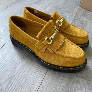 Puede incluir: Mocasines de ante amarillo mostaza con hebilla dorada y flecos. Los zapatos tienen una suela negra gruesa con costuras amarillas. Los mocasines se exhiben sobre una superficie de madera clara.