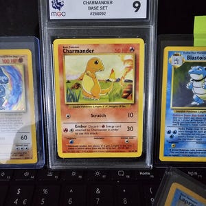 May include: A graded 1999 Pokémon Charizard Base Set trading card, featuring a vibrant image of the fire-type Pokémon. The card is encased in a protective holder with a grade of 9. Other Pokémon cards are visible in the background.