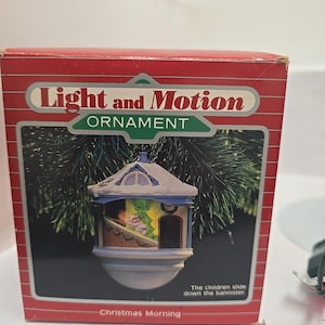 May include: A red box with a Christmas ornament inside. The ornament is a small, detailed house with a snowy roof and a scene of children sliding down a bannister. The box reads "Light and Motion Ornament" and "Christmas Morning."
