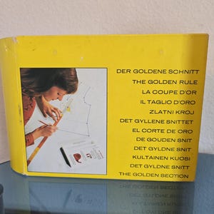 May include: A yellow rectangular instructional book titled The Golden Rule, featuring a cover image of a person using a measuring tape and pencil on a sewing pattern. The text lists the title in multiple languages including The Golden Section.
