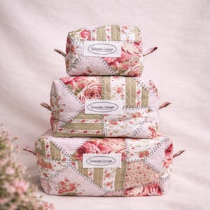 Könnte beinhalten: Ein Stapel aus drei rechteckigen, gesteppten Stofftaschen in verschiedenen Größen. Jede Tasche hat ein Patchwork-Design mit Blumenmustern in Rosa, Grün und Weiß sowie ein weißes Etikett mit der Aufschrift Keepsake Cottage in schwarzer Schrift.