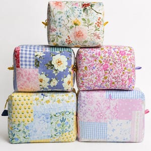 Peut inclure: Une pile de cinq trousses rectangulaires en tissu présentant divers motifs floraux et vichy en patchwork dans des tons de rose, jaune, bleu et blanc. Chaque trousse possède une fermeture éclair et une petite languette en tissu sur le côté.