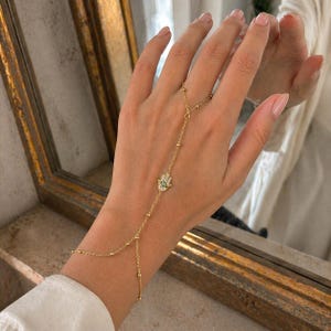 Puede incluir: Cadena de mano dorada con un dije de mano de Hamsa con una pequeña piedra turquesa en el centro. La cadena está adornada con pequeñas cuentas redondas y se conecta a un anillo. La joya se exhibe sobre un fondo neutro.