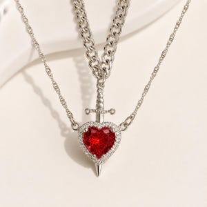 May include: A silver-toned necklace featuring a heart-shaped pendant with a vibrant red center stone, surrounded by smaller clear stones. A sword design pierces the heart. The necklace has two chains, one a delicate cable chain and the other a thicker curb chain.