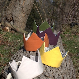 Puede incluir: Una colección de coronas de tela coloridas en tonos naranja, amarillo, morado, verde y blanco roto. Las coronas están dispuestas sobre un tronco de árbol, mostrando su diseño puntiagudo y patrones interiores. Coronas para niños.