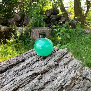 May include: A spherical glass bottle filled with a vibrant, translucent green liquid. The bottle features a smooth, rounded body and is sealed with a small, black, ribbed screw-top cap. The container is clear, allowing the bright green color to show.