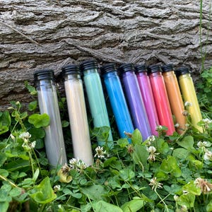 May include: A row of eight clear cylindrical glass vials with black screw-on caps, each filled with a different colored substance. The colors include grey, cream, mint green, bright blue, purple, magenta, pink, orange, and yellow.