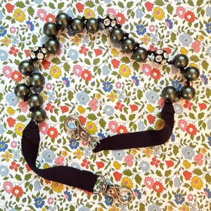 May include: A decorative strap featuring a series of dark olive green spherical beads interspersed with three round silver-toned beads encrusted with clear crystals. The beaded section connects to a black ribbed fabric strap with two metal swivel clips.