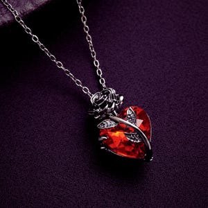 May include: A silver necklace featuring a heart-shaped, red gemstone pendant. The pendant is surrounded by a silver rose and leaves. The chain is fine, set against a dark purple backdrop.