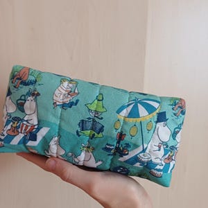 Può includere: Un astuccio rettangolare in tessuto trapuntato con sfondo verde acqua con un motivo ripetuto di personaggi dei cartoni animati. I personaggi sono bianchi con vari accessori, impegnati in attività ricreative. L'astuccio è tenuto in una mano.