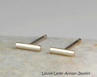 May include: A pair of gold-toned metal bar stud earrings with a smooth, polished finish. The earrings feature a horizontal rectangular bar design with a post backing. The text Louise Leder Artisan Jewelry is printed in black on the surface.