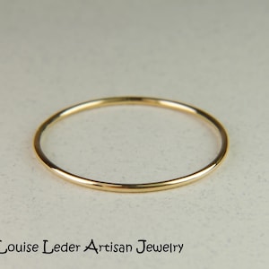 May include: A thin, circular gold ring with a smooth, polished finish. The band is uniform in thickness and features a reflective metallic surface. The text 'Louise Leder Artisan Jewelry' is printed in black lettering below the ring.