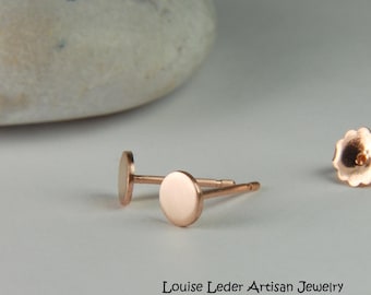 May include: Rose gold stud earrings with a simple, minimalist design. The earrings have a small, flat, circular shape. The image also shows the earring backs. The text "Louise Leder Artisan Jewelry" is visible at the bottom of the image.