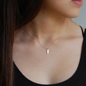 May include: A delicate gold necklace featuring a small, rectangular pendant. The pendant hangs from a fine chain, creating a minimalist and elegant look. The necklace is worn against a black top, highlighting its subtle shine.