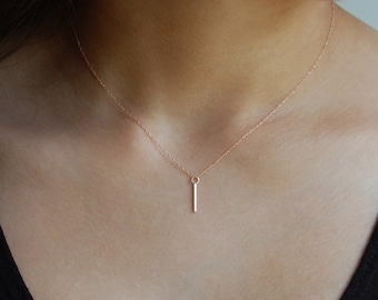May include: A delicate rose gold necklace with a slender vertical bar pendant. The pendant hangs from a fine chain, creating a minimalist design. The necklace is shown against a black top.