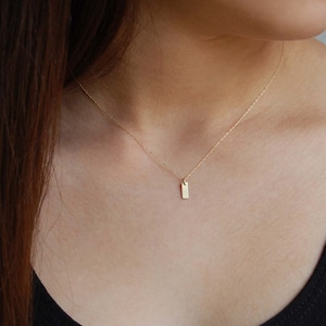 May include: A delicate gold necklace with a small, rectangular pendant. The pendant hangs from a fine chain, creating a minimalist and elegant look. The necklace is shown against a dark background.