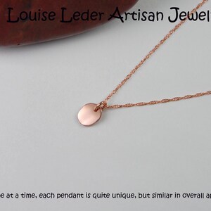 Puede incluir: Un collar de oro rosa con un peque&ntilde;o colgante redondo. La fina cadena se muestra sobre un fondo blanco. El texto "Louise Leder Artisan Jewelry" es visible en la parte superior. El texto en la parte inferior dice: "Hecho uno a la vez..."