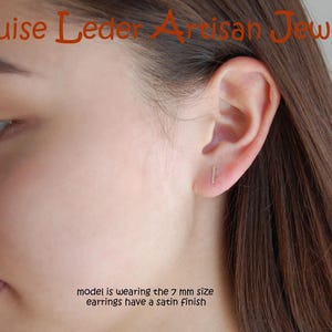 May include: A close-up of a person's face and ear, showcasing a small, gold-colored bar earring. The earrings have a satin finish. The text "Louise Leder Artisan Jewelry" is at the top. The text "model is wearing the 7 mm size earrings have a satin finish" is at the bottom.