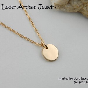 May include: A gold-coloured necklace featuring a small, circular pendant. The fine chain supports a polished, disc-shaped charm. The text "Louise Leder Artisan Jewelry" is at the top. The text "Minimalist. And just a bit rustic. Perfect for everyday." is at the bottom.