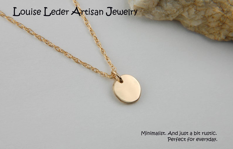 May include: A gold-coloured necklace featuring a small, circular pendant. The fine chain supports a polished, disc-shaped charm. The text "Louise Leder Artisan Jewelry" is at the top. The text "Minimalist. And just a bit rustic. Perfect for everyday." is at the bottom.
