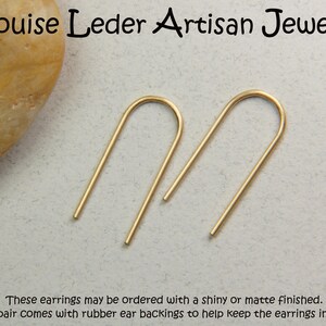 May include: A pair of gold-toned U-shaped wire earrings. The text at the top reads Louise Leder Artisan Jewelry, and the text at the bottom reads These earrings may be ordered with a shiny or matte finished. Each pair comes with rubber ear backings.