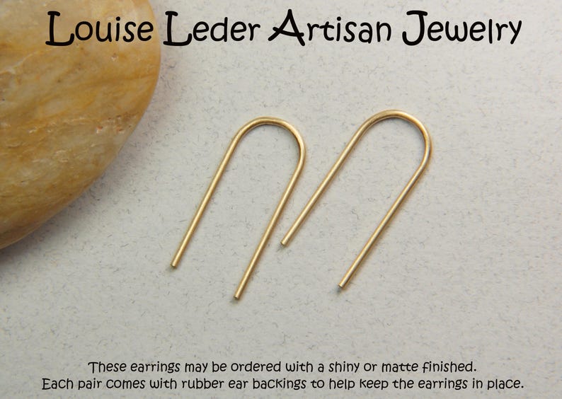 May include: A pair of gold-toned U-shaped wire earrings. The text at the top reads Louise Leder Artisan Jewelry, and the text at the bottom reads These earrings may be ordered with a shiny or matte finished. Each pair comes with rubber ear backings.