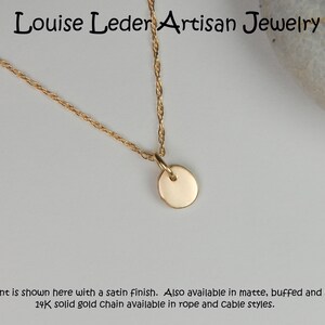 May include: A gold-colored necklace with a small, round pendant. The chain is a delicate rope style. The text "Louise Leder Artisan Jewelry" is at the top. The pendant is shown with a satin finish.