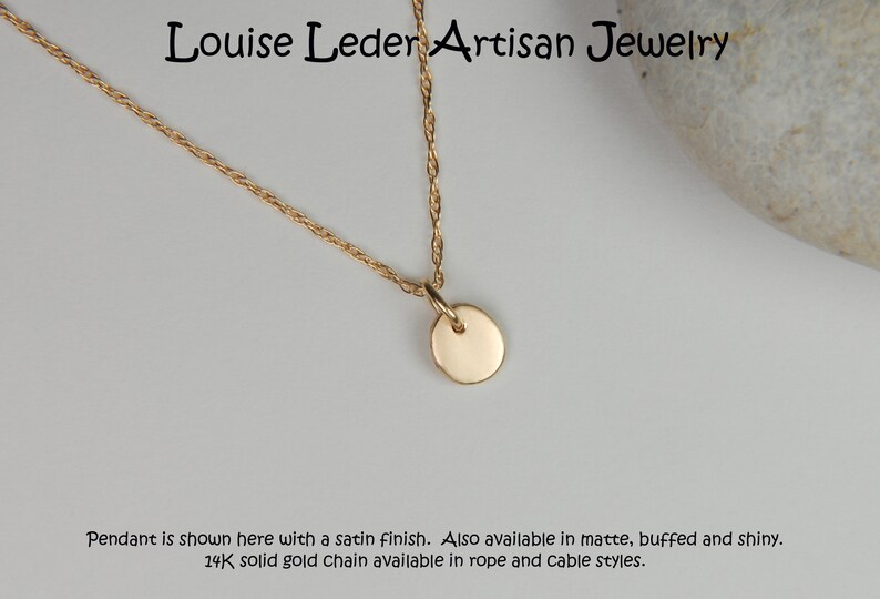 May include: A gold-colored necklace with a small, round pendant. The chain is a delicate rope style. The text "Louise Leder Artisan Jewelry" is at the top. The pendant is shown with a satin finish.