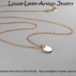 May include: A gold-coloured necklace featuring a small, oval pendant. The chain is finely linked, and the pendant has a shiny finish. The text "Louise Leder Artisan Jewelry" is at the top. Also available in matte, satin and buffed.