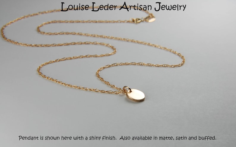 May include: A gold-coloured necklace featuring a small, oval pendant. The chain is finely linked, and the pendant has a shiny finish. The text "Louise Leder Artisan Jewelry" is at the top. Also available in matte, satin and buffed.