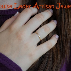 May include: A thin, polished gold band ring worn on a finger. The text "Louise Leder Artisan Jewelry" is displayed in orange lettering at the top of the image. The ring features a smooth, minimalist circular design with a reflective metallic finish.