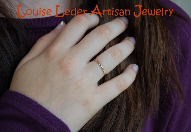 May include: A thin, polished gold band ring worn on a finger. The text "Louise Leder Artisan Jewelry" is displayed in orange lettering at the top of the image. The ring features a smooth, minimalist circular design with a reflective metallic finish.