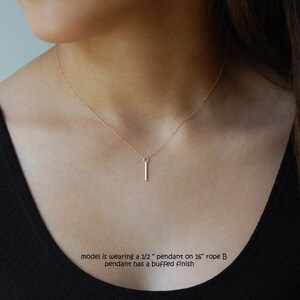 Puede incluir: Un delicado collar de oro rosa con un colgante de barra vertical. El colgante mide aproximadamente 1,27 cm de largo y cuelga de una cadena de 40,64 cm. El colgante tiene un acabado pulido. El collar se lleva sobre un top negro.