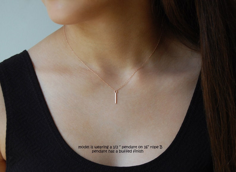 Puede incluir: Un delicado collar de oro rosa con un colgante de barra vertical. El colgante mide aproximadamente 1,27 cm de largo y cuelga de una cadena de 40,64 cm. El colgante tiene un acabado pulido. El collar se lleva sobre un top negro.