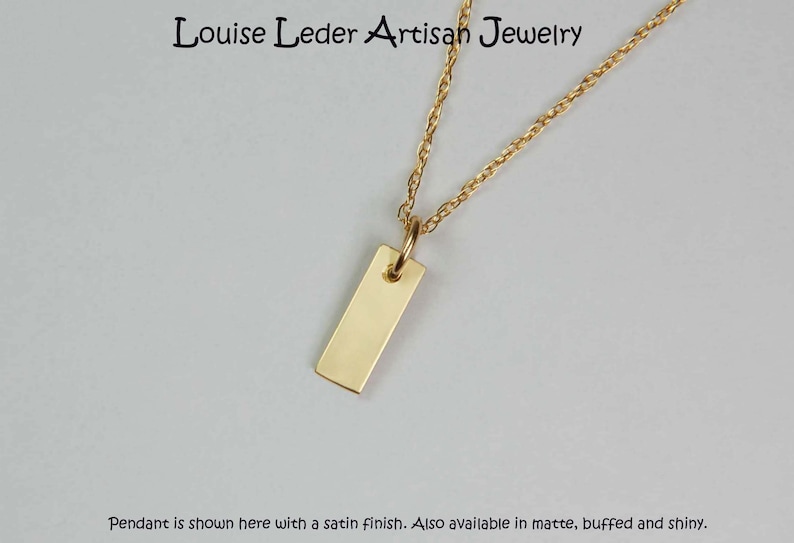 May include: A gold-coloured rectangular pendant necklace with a delicate chain. The pendant is a simple, polished rectangle. The text "Louise Leder Artisan Jewelry" is at the top. The pendant is shown with a satin finish.