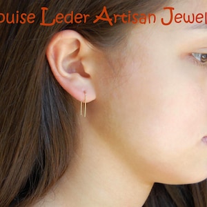 May include: A gold-toned U-shaped wire earring worn in the earlobe. The text Louise Leder Artisan Jewelry is visible at the top of the image. The earring features a simple, thin, rectangular wire design that hangs vertically from the ear piercing.