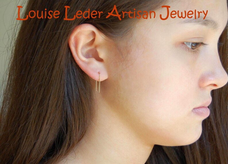May include: A gold-toned U-shaped wire earring worn in the earlobe. The text Louise Leder Artisan Jewelry is visible at the top of the image. The earring features a simple, thin, rectangular wire design that hangs vertically from the ear piercing.