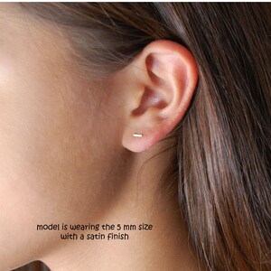 Puede incluir: Primer plano de la oreja de una persona con un peque&ntilde;o pendiente de barra de oro delgado. La modelo lleva la talla de 5 mm con un acabado satinado.