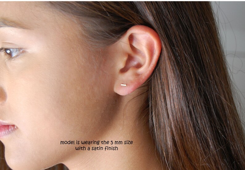 Puede incluir: Primer plano de la oreja de una persona con un peque&ntilde;o pendiente de barra de oro delgado. La modelo lleva la talla de 5 mm con un acabado satinado.