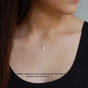 May include: A gold necklace featuring a small, rectangular pendant. The pendant, about 8 mm, hangs from a 16-inch rope chain. The pendant has a satin finish. The model is wearing a black top.