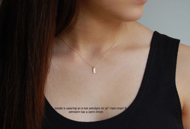 May include: A gold necklace featuring a small, rectangular pendant. The pendant, about 8 mm, hangs from a 16-inch rope chain. The pendant has a satin finish. The model is wearing a black top.