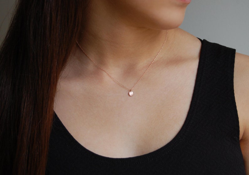 Puede incluir: Un delicado collar de oro rosa con un peque&ntilde;o colgante redondo. El colgante cuelga de una fina cadena, creando un look minimalista y elegante. El collar se lleva sobre una camiseta negra, resaltando su sutil brillo.