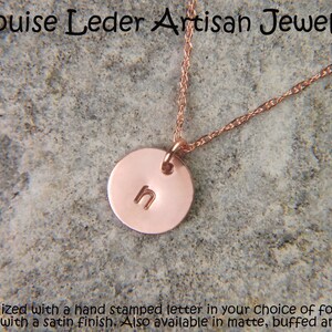 May include: A rose gold-toned circular pendant necklace featuring the hand-stamped lowercase letter n. The pendant hangs from a delicate twisted chain. The text Louise Leder Artisan Jewelry is visible at the top, with descriptive text below.