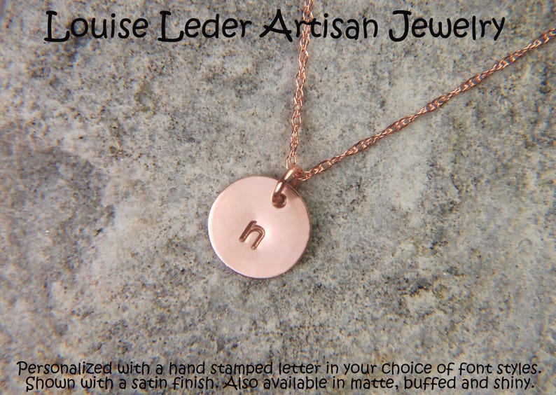 May include: A rose gold-toned circular pendant necklace featuring the hand-stamped lowercase letter n. The pendant hangs from a delicate twisted chain. The text Louise Leder Artisan Jewelry is visible at the top, with descriptive text below.