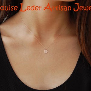 May include: A delicate rose gold necklace featuring a small, circular pendant with a single engraved letter. The text Louise Leder Artisan Jewelry is displayed in orange at the top of the image. The necklace rests against a black sleeveless top.
